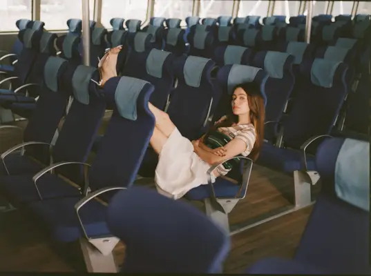 Molly Gordon sitting in a sea of empty chairs on a ferry boat. She has her arms crossed and her feet perched up on the chair in front of her. She is wearing a striped Miu Miu shirt and white skirt.