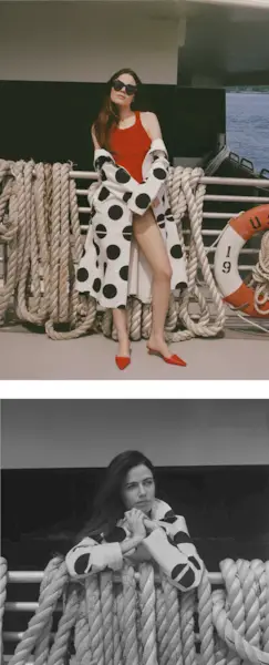 Molly Gordon standing outside on a ferry boat. In the top image, she is standing in front of railing with large ropes hanging over it. She is wearing a polka dot coat off the shoulders with a red one-piece bathing suit and red heels. In the bottom image, she is leaning over the railing with the ropes looking off camera.