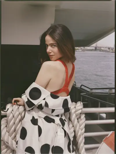 Molly Gordon standing on the outside of a ferry boat. She is looking over her shoulder wearing a red one-piece Tory Burch bathing suit with a black and white polka dot Renaissance Renaissance jacket hanging off her shoulders.