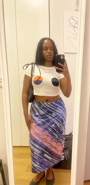 A woman wearing a colorful sequin skirt, white top with colorful circles printed, a black bag, and black flats