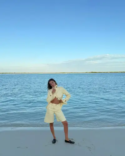 Sandra wearing a yellow jacket and matching Bermuda shorts with black loafers on a white sand beach.