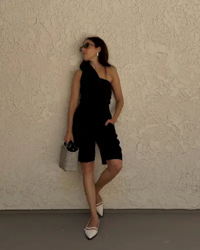 Lauren wearing a one-shoulder black top, black Bermuda shorts, and white slingback flats in front of a beige wall.