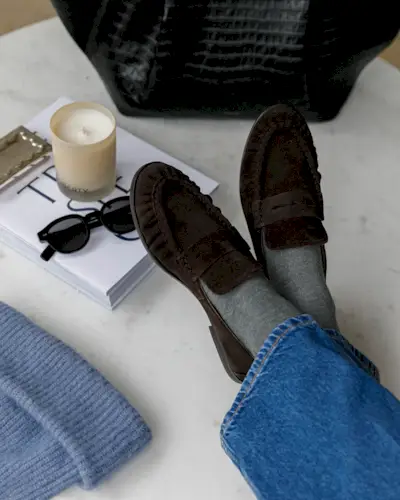 Fashion person Kim Turkington wears brown shoe outfits. In the recent image taken from her Instagram she has brown suede loafers on a marble table. Around her is a white coffee table book a candle and a black croc bag.