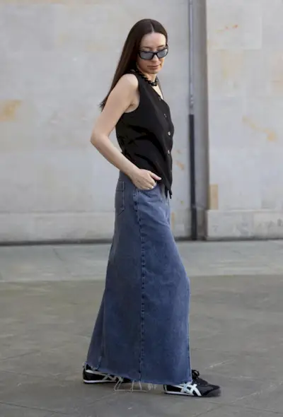 A woman wearing a long denim skirt with sneakers and black waistcoat.