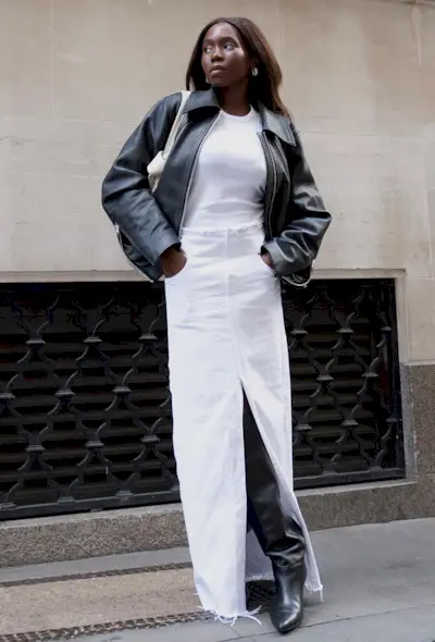 A woman wearing a long white denim skirt with a white T-shirt, a leather bomber jacket, and knee-high boots