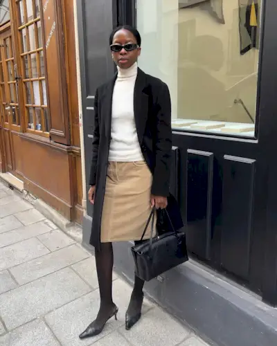 sylvie wears a black coat, white turtleneck, long black jacket, sheer black tights and black slingback heels.