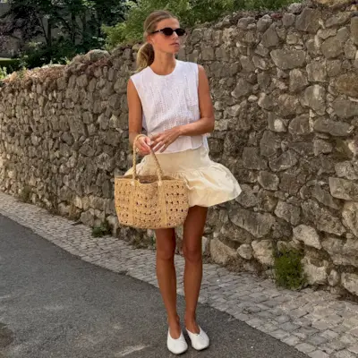 A woman wears a white top, peplum skirt, raffia bag, and white ballet flats.