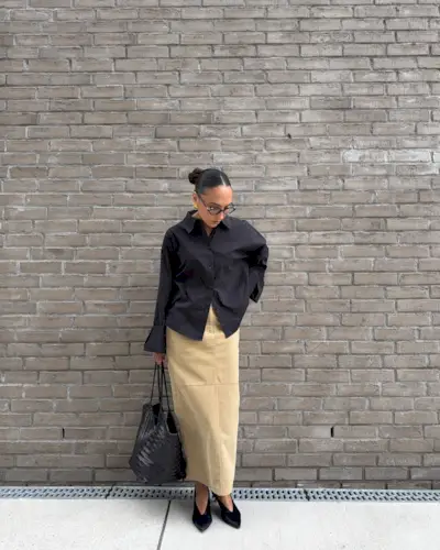 Woman wears long skirt, black shirt, pointed to heels and woven leather bag
