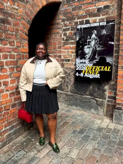 Woman wears barn jacket, white t-shirt, plaid skirt, loafers and red bag