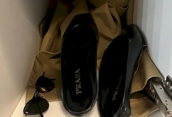 A pair of black Prada flat mules in a locker with sunglasses and a trench coat.