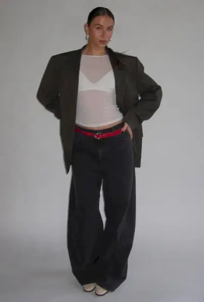 A woman standing inside a white studio space wearing an oversize blazer over a white sheer t-shirt and white bralette styled with a red belt, black baggy jeans, silver earrings, and white loafers.