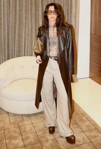 A woman standing by a white armchair wearing a dark brown leather trench coat over a brown printed mesh t-shirt styled with a dark skinny belt, brown clutch, light tan wide leg trousers, and dark brown platform ankle boots.