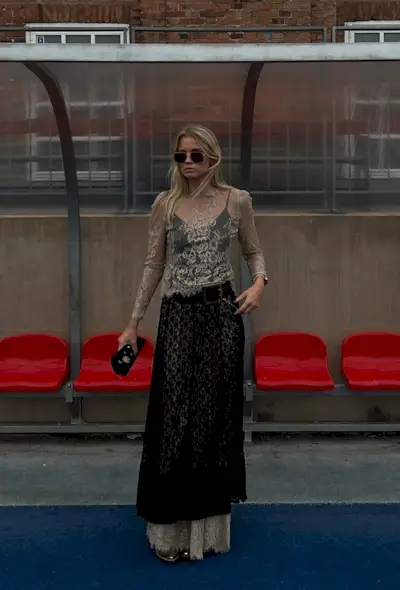 A woman standing outside on a track field wearing a light tan sheer lace shirt layered over a black sheer lace dress layered over a tan sheer lace skirt, gold flats, a black belt, gold bangles, aviator sunglasses, and a black clutch bag.
