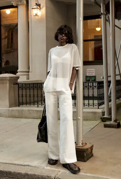 A woman standing outside on the sidewalk wearing a white oversize sheer t-shirt over a white satin bra with white linen wide-leg trousers, black flat sandals, a black tote bag, gold bangles, and black sunglasses.