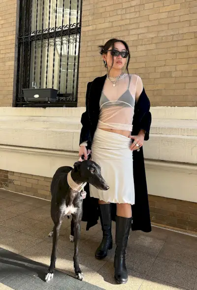 A woman standing outside with her dog wearing a long black coat over a white sheer top styled with a black bralette, silver chunky chain, white balloon midi skirt, stacked silver rings, black sporty sunglasses, and black knee-high heeled boots.