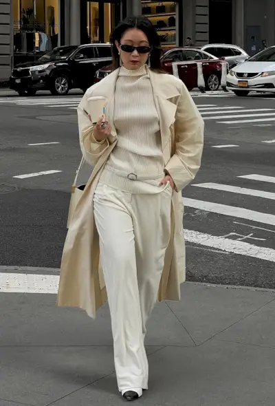 A woman walking on the sidewalk in New York wearing a white trench coat over a white pleated sheer high-neck top with a skinny white belt, white wide leg trousers, a white handbag, and white cap toe slingback heels.