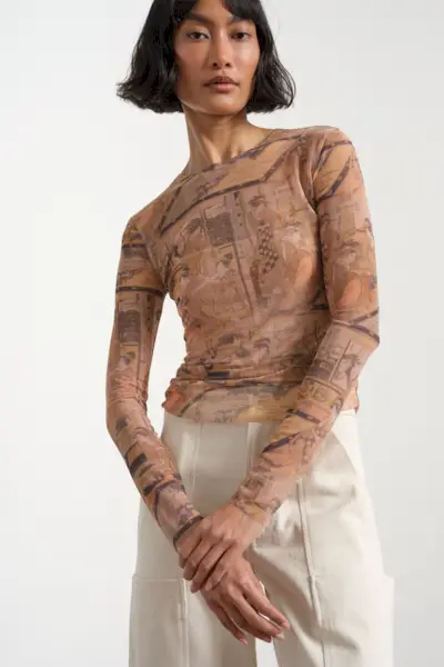 A woman walking across the crosswalk wearing a long black coat over a black sheer turtleneck and gray cardigan styled with a long silver pendant necklace, baggy white jeans, black square-toe ankle boots, and a black barrel shoulder bag.