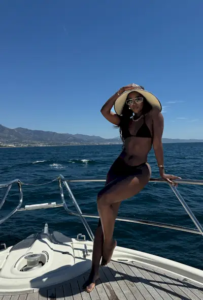 a photo showing what to wear on a boat with a woman wearing an oversized raffia hat with sunglasses, a gold chain, and a black bikini
