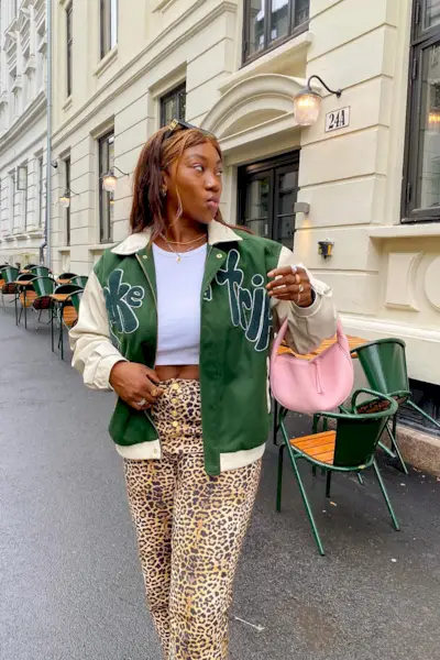 A woman standing on the sidewalk wearing a green bomber jacket over a cropped white tank top, cheetah print pants, and pink crescent bag.