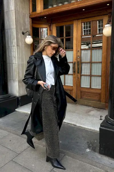 A woman walking down the sidewalk wearing a black leather trench coat over a white t-shirt, dark wash animal print pants, and black pointed ankle boots.
