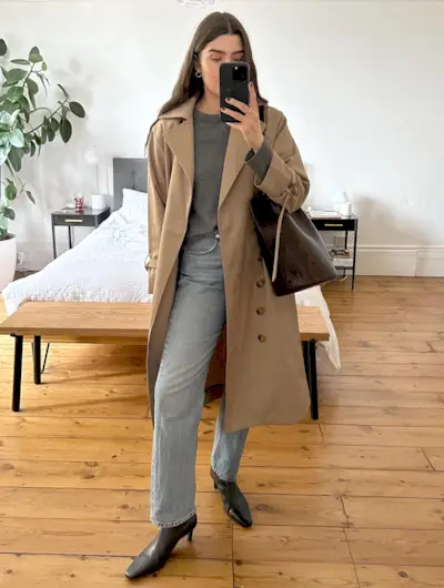 British fashion influencer Anna Newton poses for a mirror selfie wearing a trench coat, gray sweater, straight-leg jeans, and square-toe kitten heel boots.