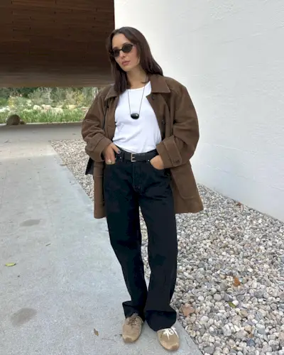 Fashion influencer Debora Rosa wearing black oval sunglasses, a black pendant necklace, white T-shirt, brown suede jacket, black belt, baggy black jeans, and Miu Miu brown suede sneakers.