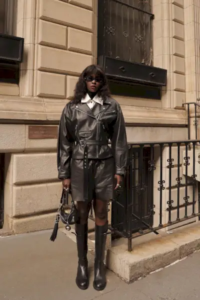 riding boot outfit shown in a photo of a woman in the street wearing a black leather moto jacket over a white button-down shirt with long black leather shorts and black knee-high riding boots