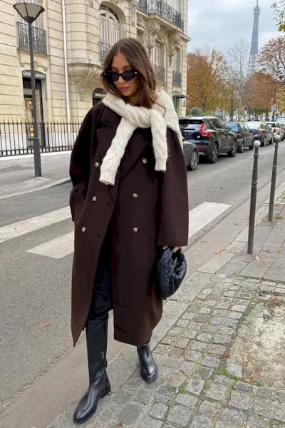 riding boot outfit shown in a photo of a woman in the street wearing a white sweater tied over a chocolate brown coat with black skinny jeans, black riding boots, and a black handbag