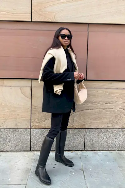 riding boot outfit shown in a photo of a woman in the street wearing a tan sweater over a black blazer with black skinny jeans and black knee high riding boots and a tan handbag