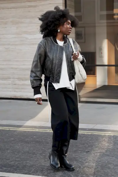 riding boot outfit shown in a photo of a woman in the street wearing a black leather bomber jacket over a white sweater with black cropped capris styled with black riding boots and a white sling bag