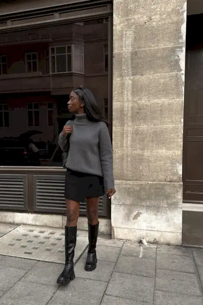 riding boot outfit shown in a photo of a woman in the street wearing a gray turtleneck sweater with a black mini skirt and black knee-high riding boots
