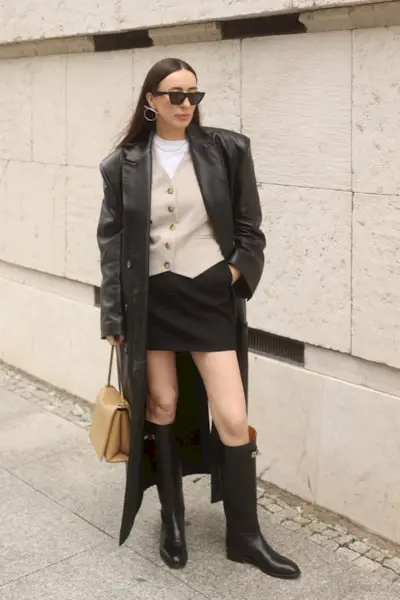 riding boot outfit shown in a photo of a woman in the street wearing a long leather trench over a white t-shirt, tan vest, and black mini skirt styled with black knee-high riding boots and a tan shoulder bag