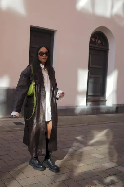 riding boot outfit shown in a photo of a woman in the street wearing a brown leather coat over a white button-down shirt dress styled with black riding boots and a green shoulder bag
