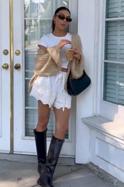 riding boot outfit shown in a photo of a woman standing in a doorway wearing brown sunglasses, a tan cardigan, a white t-shirt, white shorts, a brown bag, and brown knee-high boots