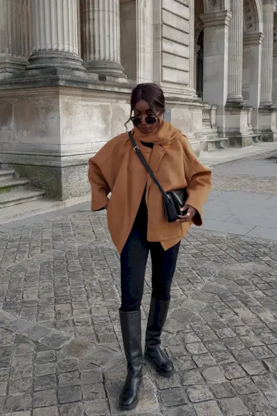 riding boot outfit shown in a photo of a woman in the street wearing a tan cropped wool coat with a built-in scarf styled with a black crossbody bag, black leggings, and black knee-high riding boots