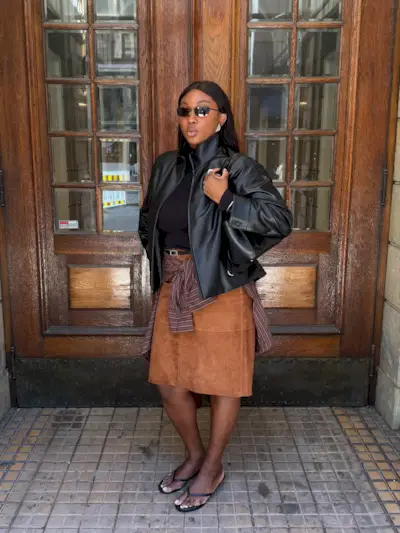 @nnennaechem wears funnel neck jacket, with a black t-shirt, striped button down shirt, flip flops and a suede midi skirt.