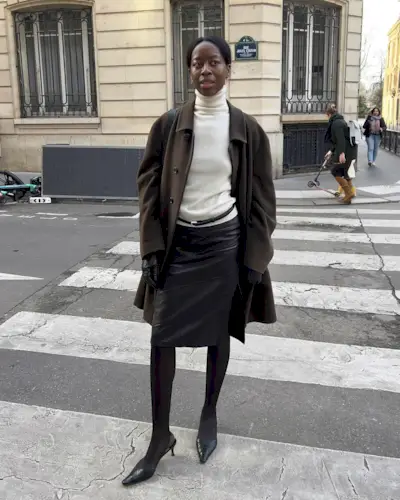@sylviemus_ wears a white turtleneck, with a brown coat, black column skirt and kitten heels.