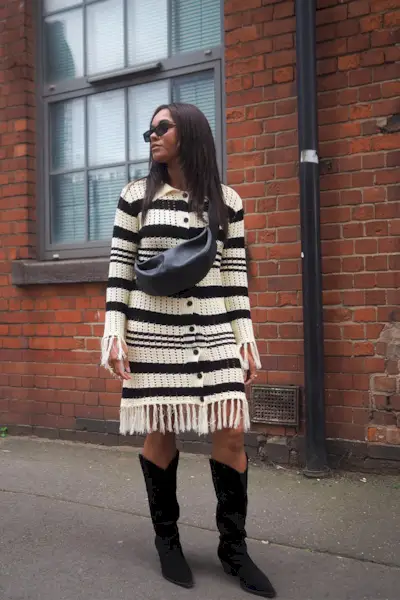 a photo of a sweater dress outfit with a woman wearing a black and white fringe sweater dress with black knee-high cowboy boots and a black sling bag