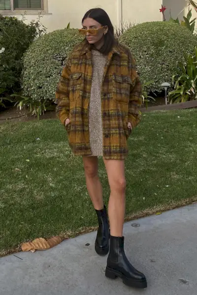 a photo of a sweater dress outfit with a woman wearing a plaid fleece jacket layered over a crewneck sweaterdress and black chelsea boots