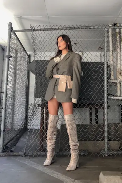 a photo of a thigh-high boots outfit shown on a woman in a park garage wearing a gray blazer over a creme sweater dress styled with snakeskin thigh-high boots