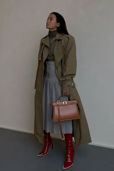 a photo of a thigh-high boots outfit shown on a woman standing against a white wall wearing a green trench coat over a turtleneck styled with a gray pleated midi-skirt and red thigh-high boots