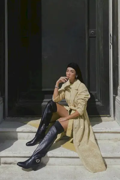a photo of a thigh-high boots outfit shown on a woman sitting on a stoop wearing an oversize yellow coat worn as a dress with black thigh-high boots