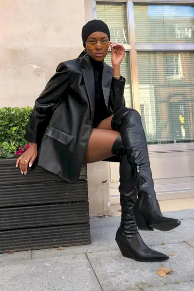 a photo of a thigh-high boots outfit shown on a woman sitting down wearing a black minidress with a black leather blazer and black thigh-high boots