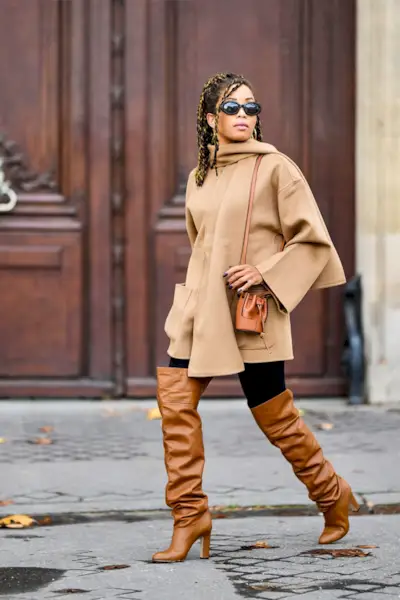 a photo of a thigh-high boots outfit shown on a woman walking down the street wearing thigh-high brown boots over black leggings with a camel wrap coat with a built-in scarf