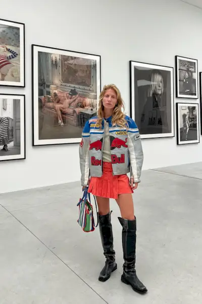 a photo of a thigh-high boots outfit shown on a woman standing in an art gallery wearing a leather moto jacket over a gray T-shirt with a red pleated miniskirt styled with black thigh-high boots