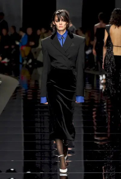 how to wear velvet is shown in a photo from Sportmax's fall 2024 runway show with a model wearing a blue button-down shirt under a black blazer tucked into a black velvet midi skirt with sheer tights and white pumps