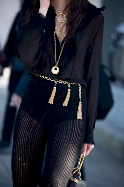 A woman wears a black blouse, tassel chain belt, and black pants.