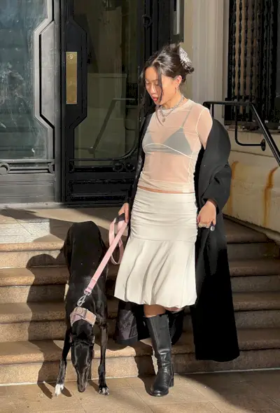 a woman wearing a white ballon skirt with a sheer white mesh top and black bra and black coat and black boots