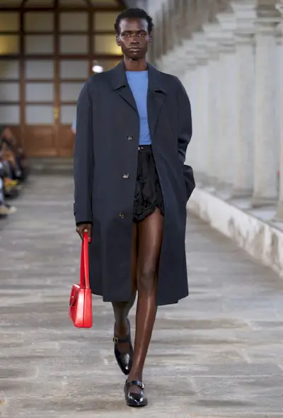 a photo of a model on Bally's spring runway wearing a black bubble skirt with a blue t-shirt and navy trench coat and black mary jane flats