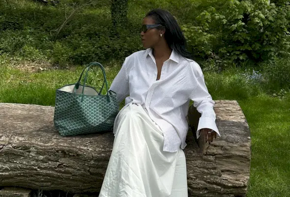 A woman wearing a button-down shirt, a poplin A-line skirt, sandals, and a green Goyard tote bag.